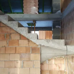 Surowe betonowe schody wewnętrzne w trakcie budowy, widoczne ceglane ściany i pustaki betonowe, elementy drewniane podtrzymujące strop.