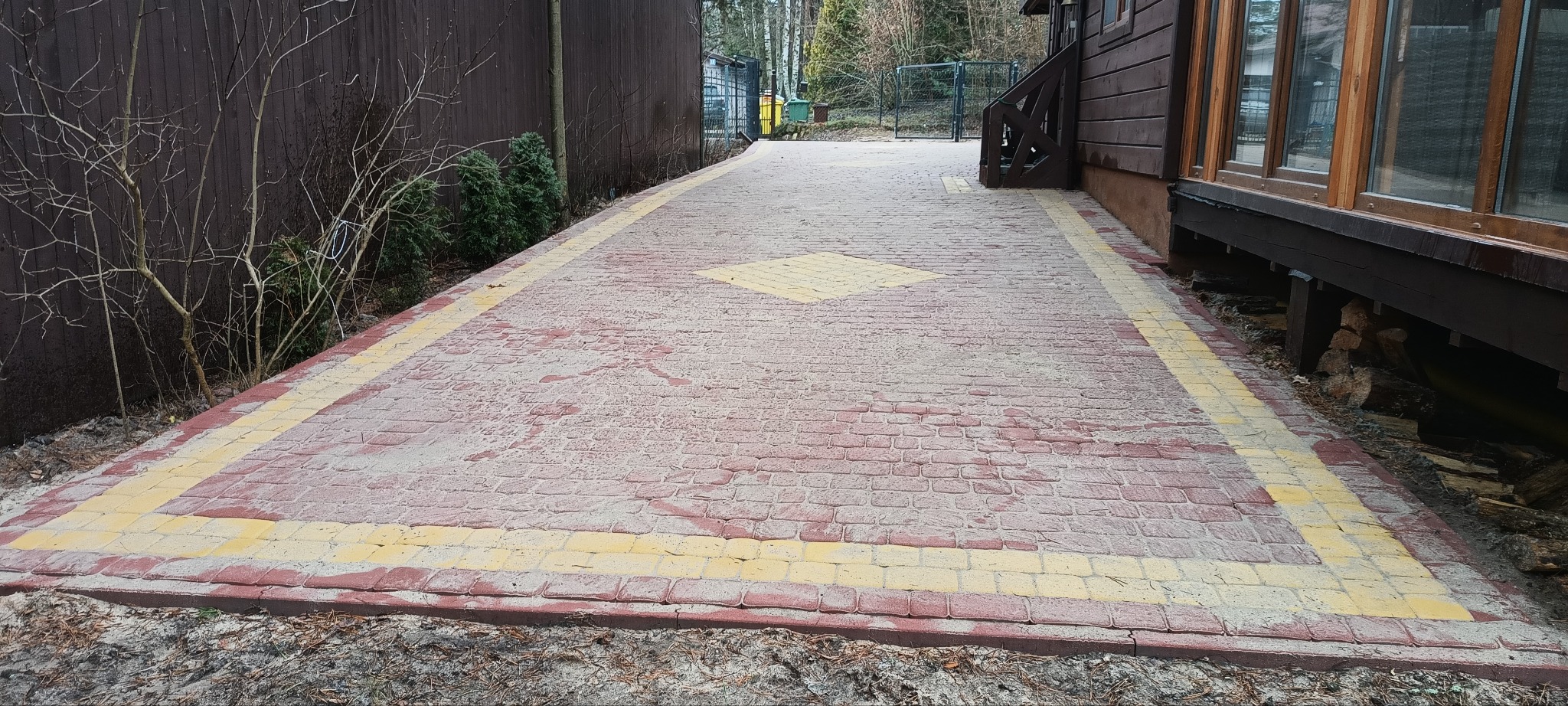 Ułożona czerwona kostka brukowa z żółtymi obramowaniami i centralnym wzorem na podwórku obok drewnianego budynku i ogrodzenia.