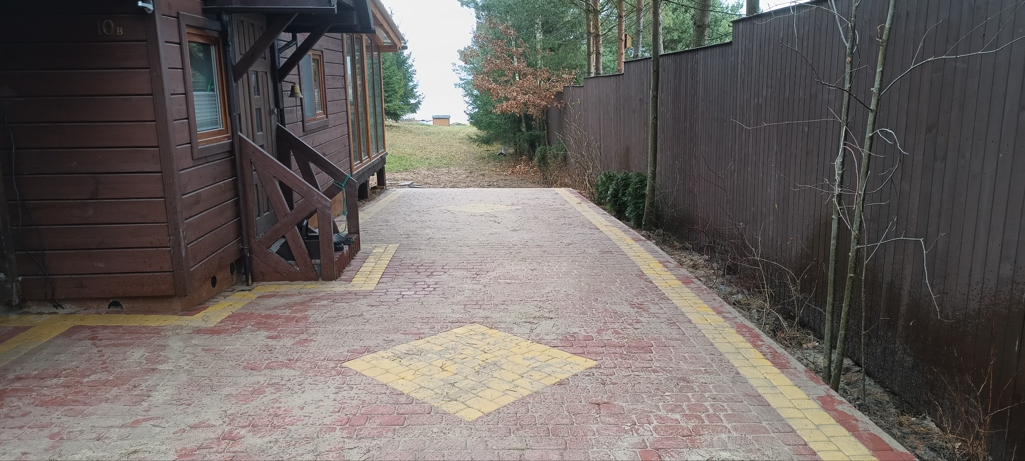 Czerwono-żółta kostka brukowa prowadząca od drewnianego domku w kierunku lasu, brązowe ogrodzenie po prawej stronie.