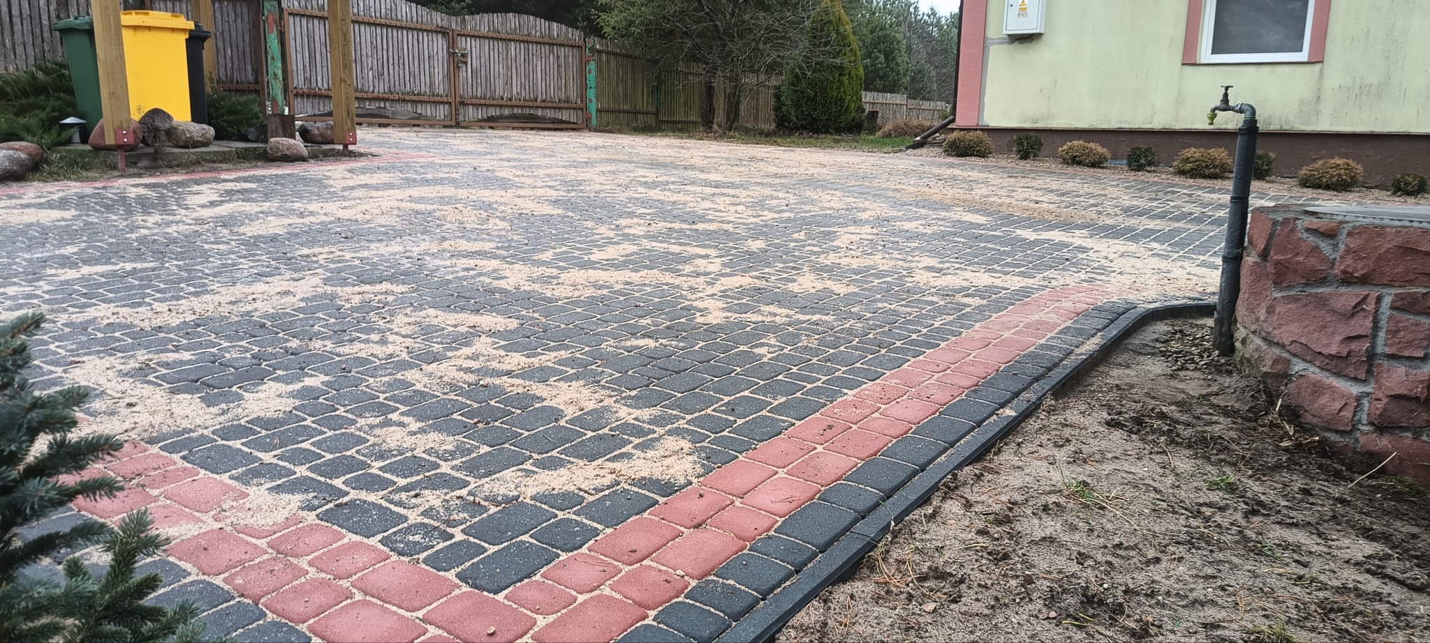 Ułożona kostka brukowa z szarych i czerwonych elementów, posypana piaskiem, widoczny fragment domu z kranem, żółty pojemnik na śmieci i ogrodzenie w tle.