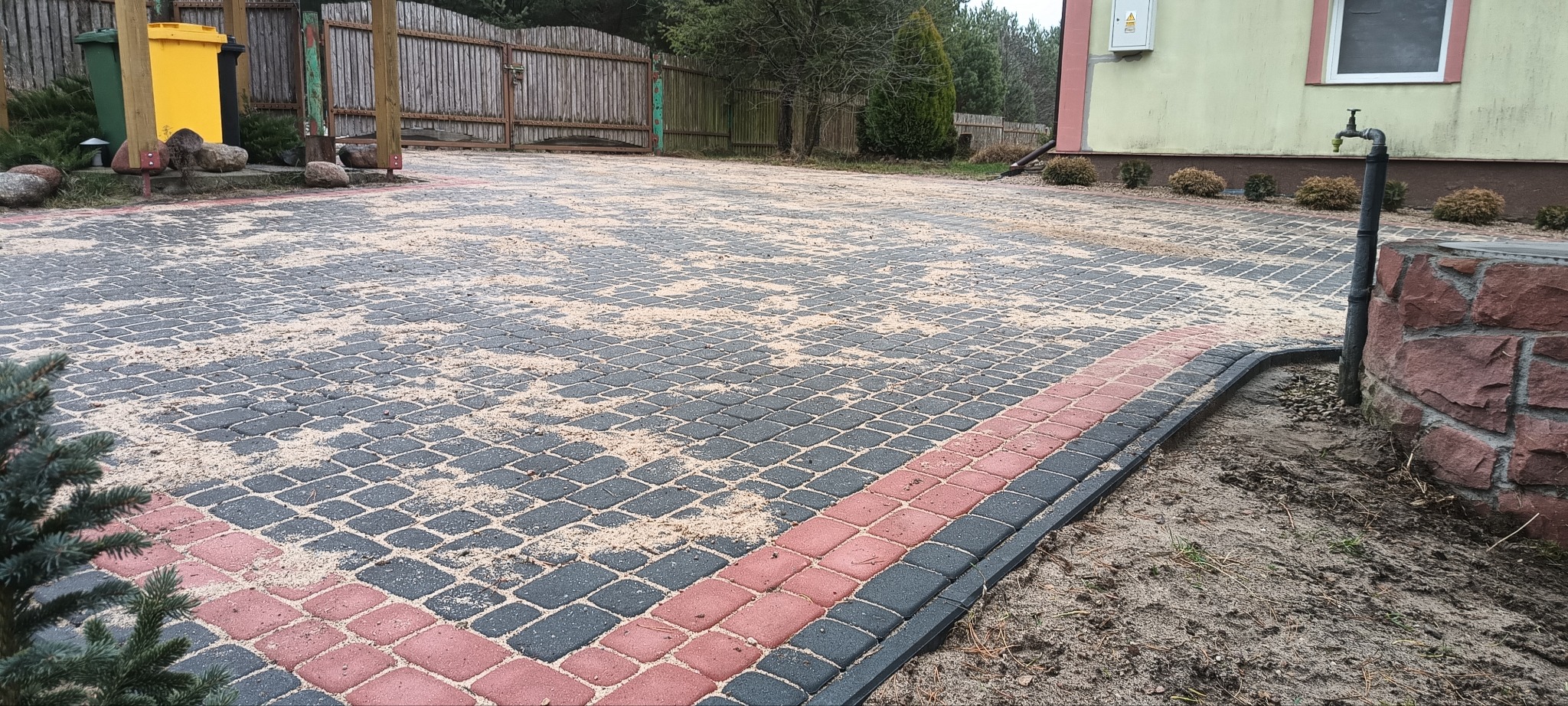 Ułożona kostka brukowa w odcieniach szarości i czerwieni, zasypana piaskiem, widok z poziomu gruntu, dom z kranem w tle.