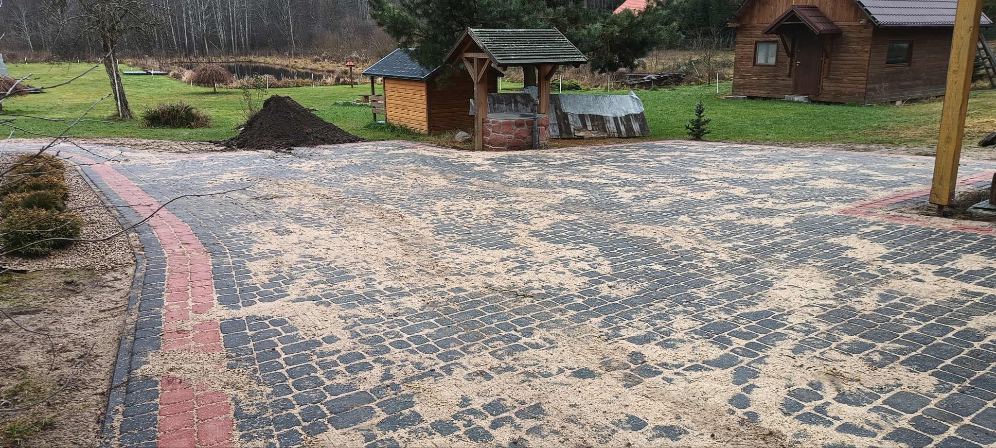 Szara kostka brukowa z czerwoną obwódką, pokryta piaskiem, na tle drewnianych budynków gospodarczych i sterty ziemi.