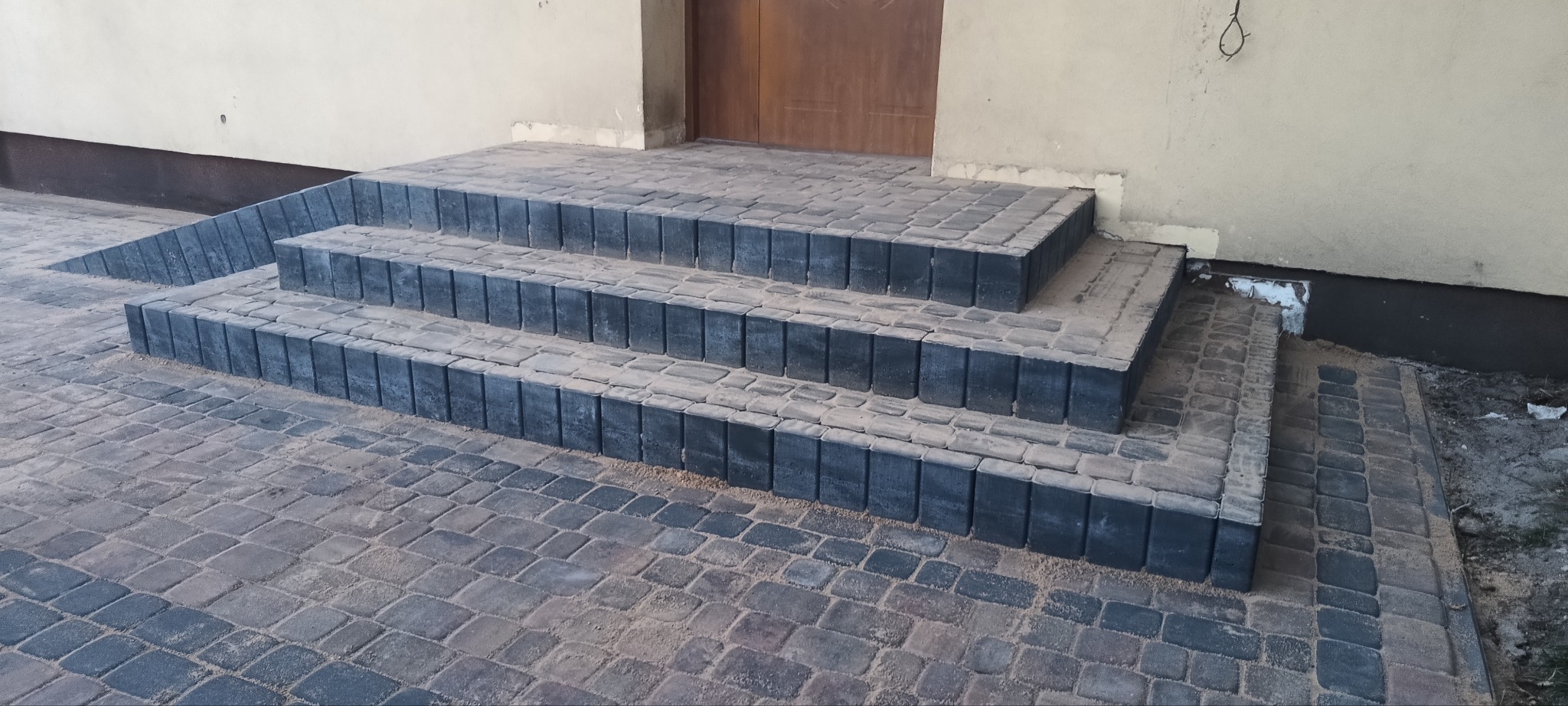 Schody wejściowe do budynku wyłożone kostką brukową w odcieniach szarości i brązu, z widoczną fugą piaskową oraz fragmentem elewacji.