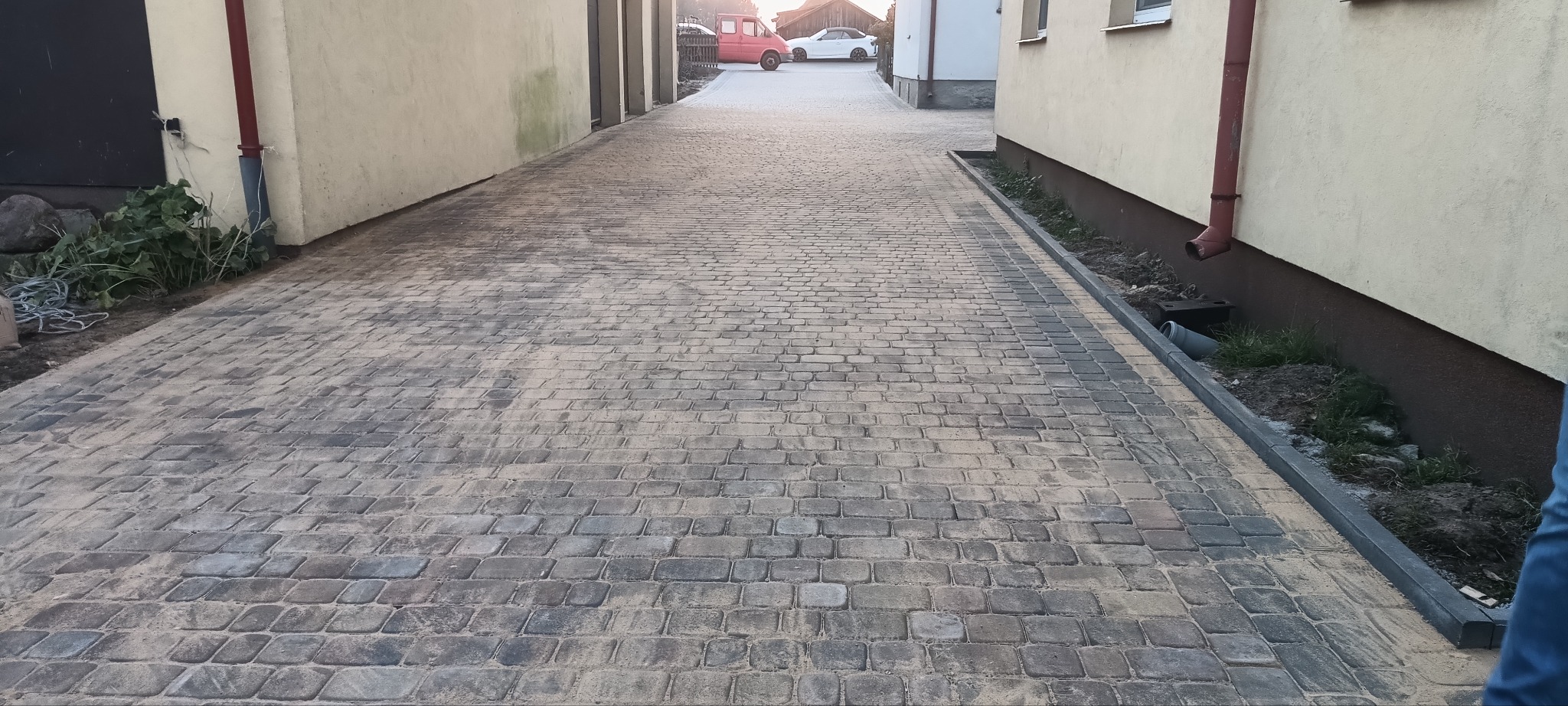 Ułożona kostka brukowa w wąskim przejściu między budynkami, widoczne krawężniki i rynna spustowa, w tle zaparkowane samochody.