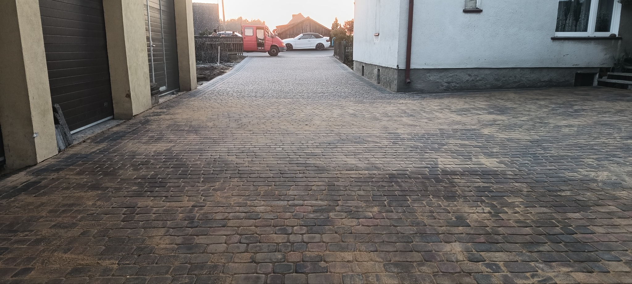 Ułożona kostka brukowa w odcieniach szarości i brązu tworzy wzór na podjeździe, widoczny budynek z białymi ścianami i garaż z brązową bramą w tle, w oddali zaparkowane samochody.