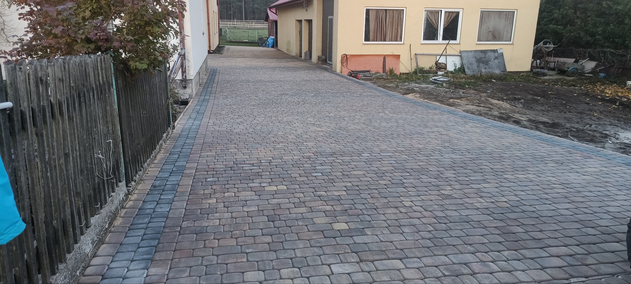 Ułożona kostka brukowa na podjeździe, obrzeża z ciemniejszej kostki, widoczne budynki w tle i fragment drewnianego płotu po lewej stronie.