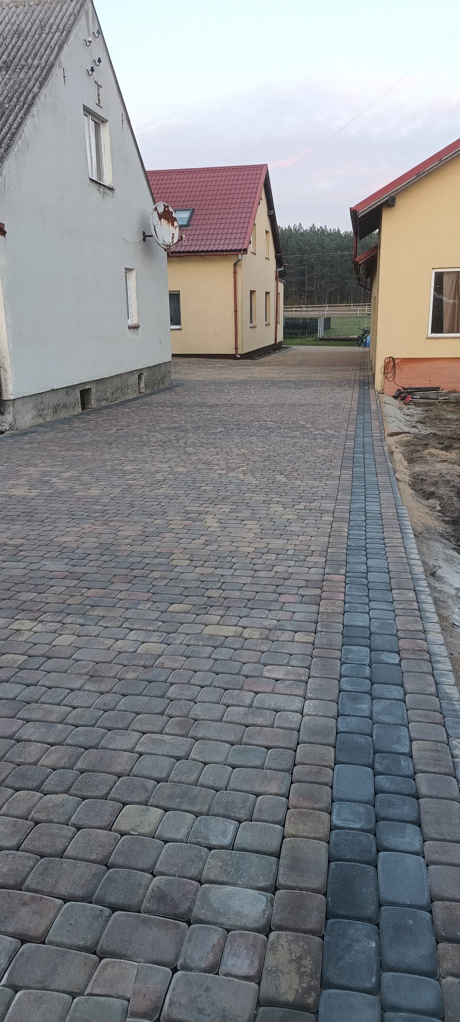 Ułożona, wielokolorowa kostka brukowa prowadząca między dwoma domami z widocznym fragmentem ogrodu w tle.