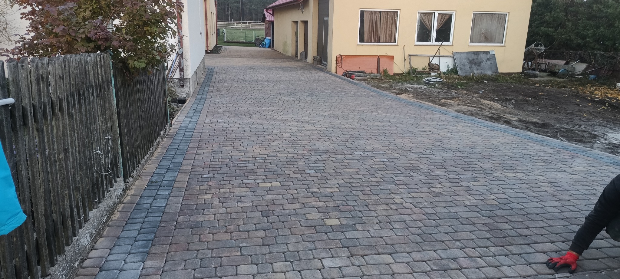 Ułożona aleja z kostki brukowej w odcieniach szarości i brązu, obramowana ciemniejszymi kostkami, prowadząca wzdłuż drewnianego płotu i budynku z żółtą elewacją.