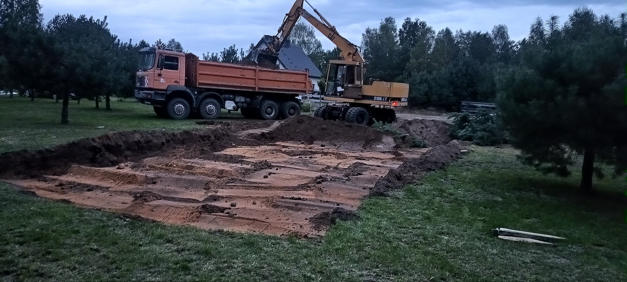 Żółta koparka CAT 212B FT ładuje ziemię do pomarańczowej wywrotki MAN, widoczny częściowo wykopany teren z warstwami piasku i ziemi, w tle drzewa i budynek mieszkalny.