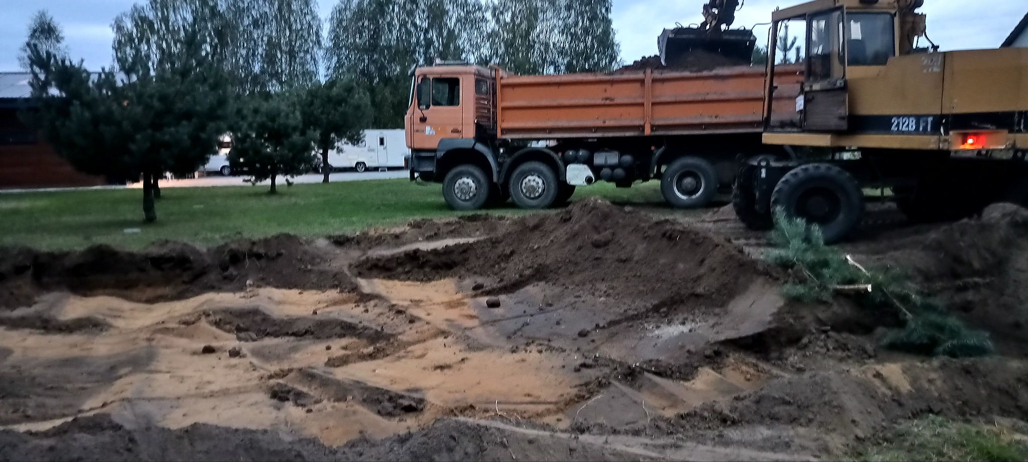 Pomarańczowa wywrotka i żółta koparka podczas prac ziemnych na działce, widoczne wykopane doły i usypana ziemia, w tle zieleń drzew i biały kamper.