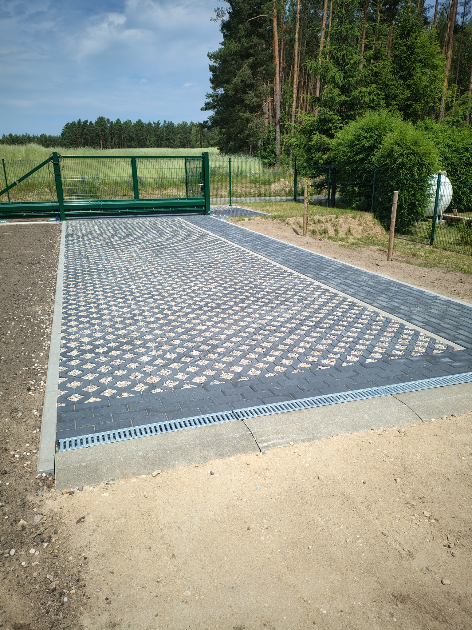 Ułożona ażurowa kostka brukowa z wypełnieniem żwirowym, obrzeża z kostki, drenaż liniowy, brama wjazdowa i ogrodzenie panelowe na tle drzew.