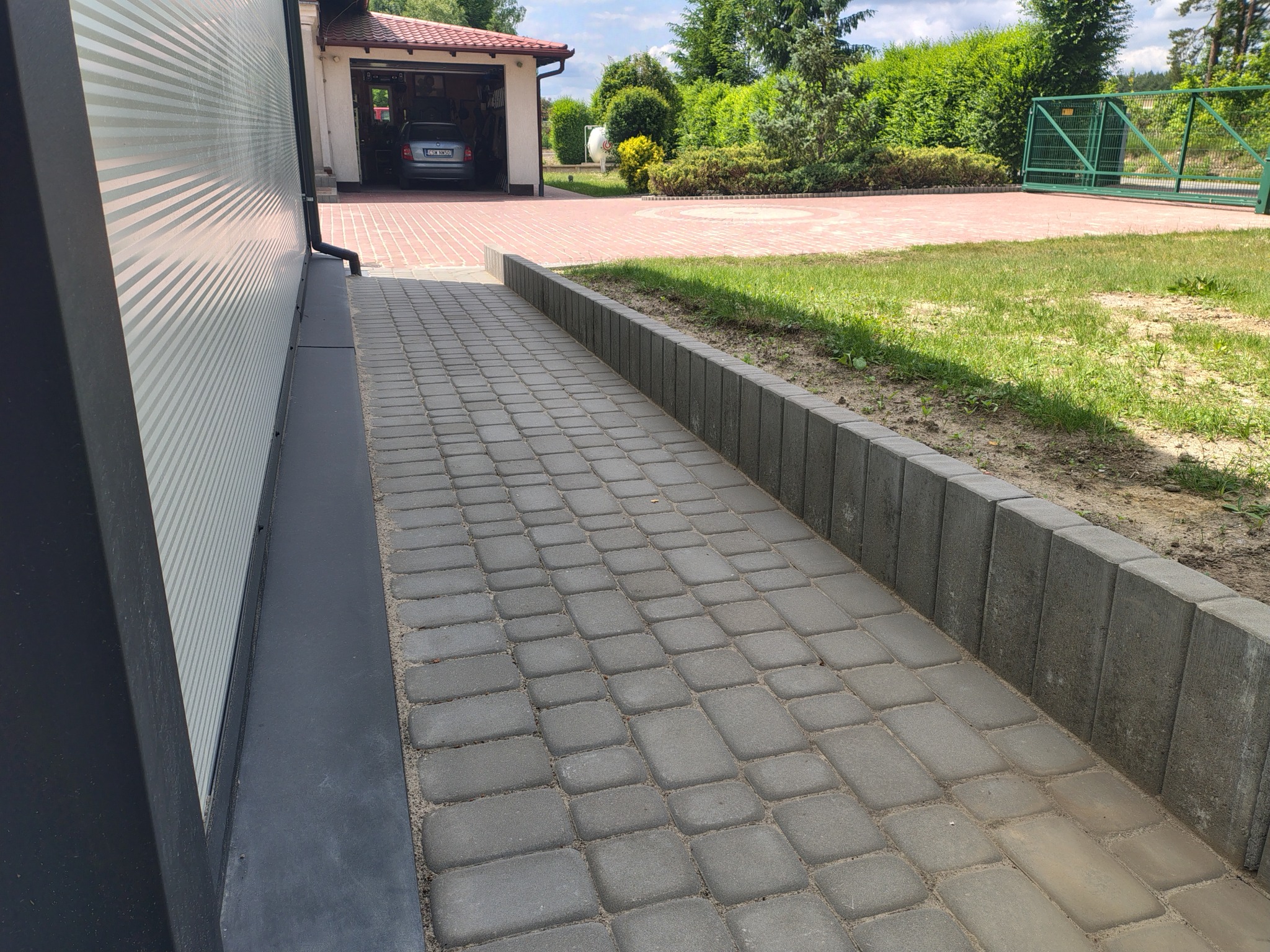 Ułożona szara kostka brukowa tworząca ścieżkę obok budynku z garażem w tle, widok na podjazd i zieleń ogrodu.