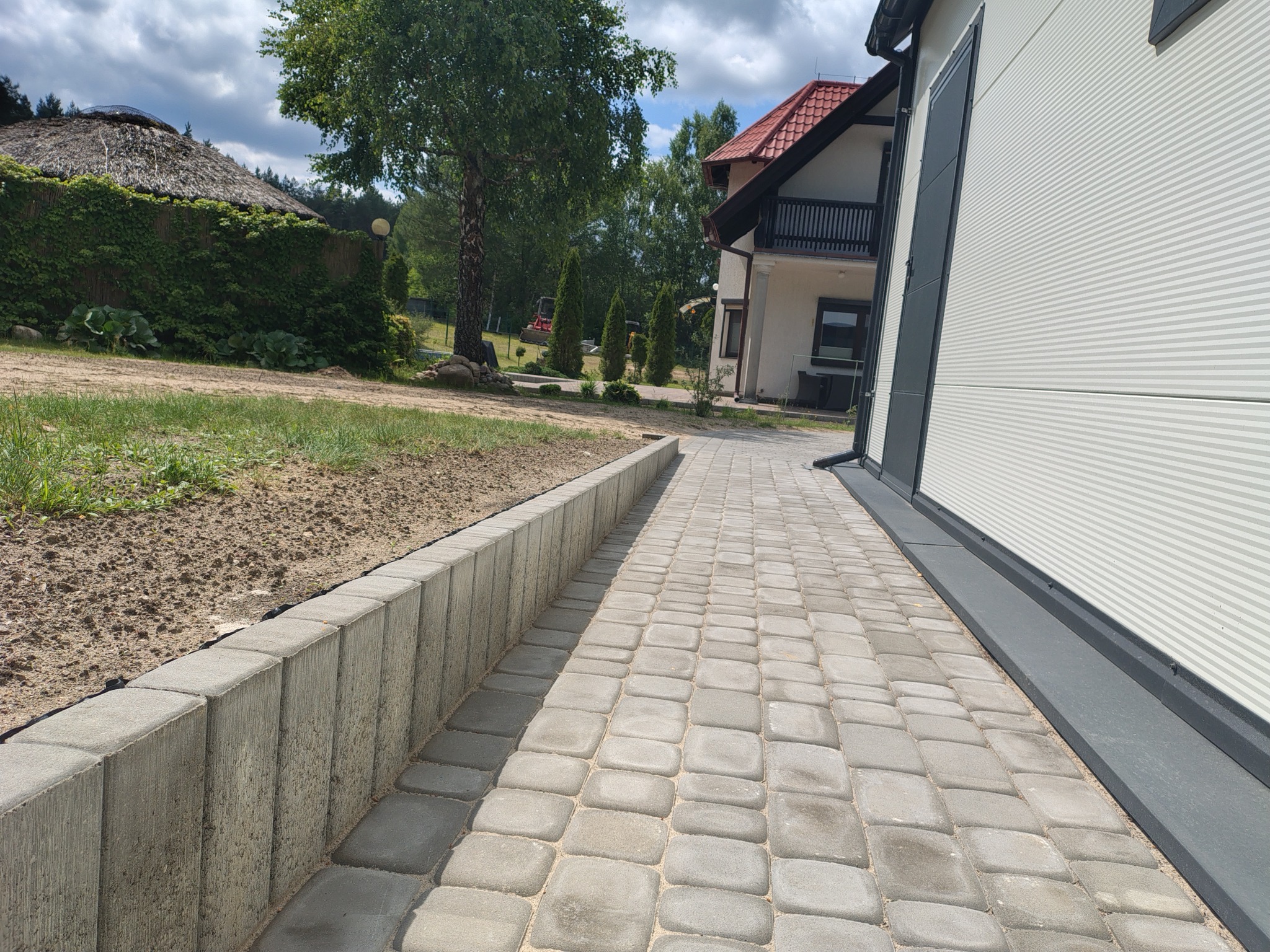 Ścieżka z szarej kostki brukowej prowadząca wzdłuż budynku z szarą elewacją, obok rabata ziemi i niski murek. W tle domek z dachem krytym strzechą.