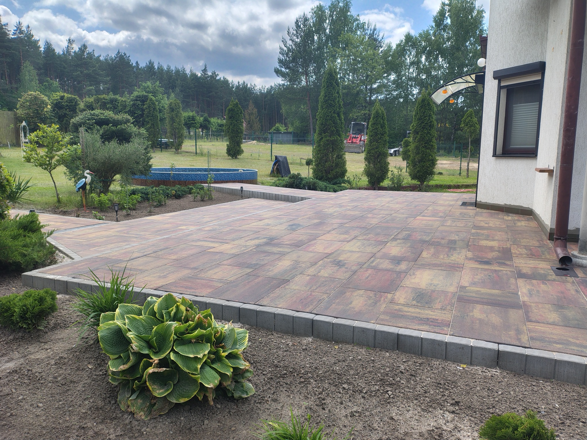 Rozległy taras z kostki brukowej w odcieniach brązu i czerwieni, otoczony zielenią i fragmentem domu z oknem. W tle widoczny basen i zadbany ogród.