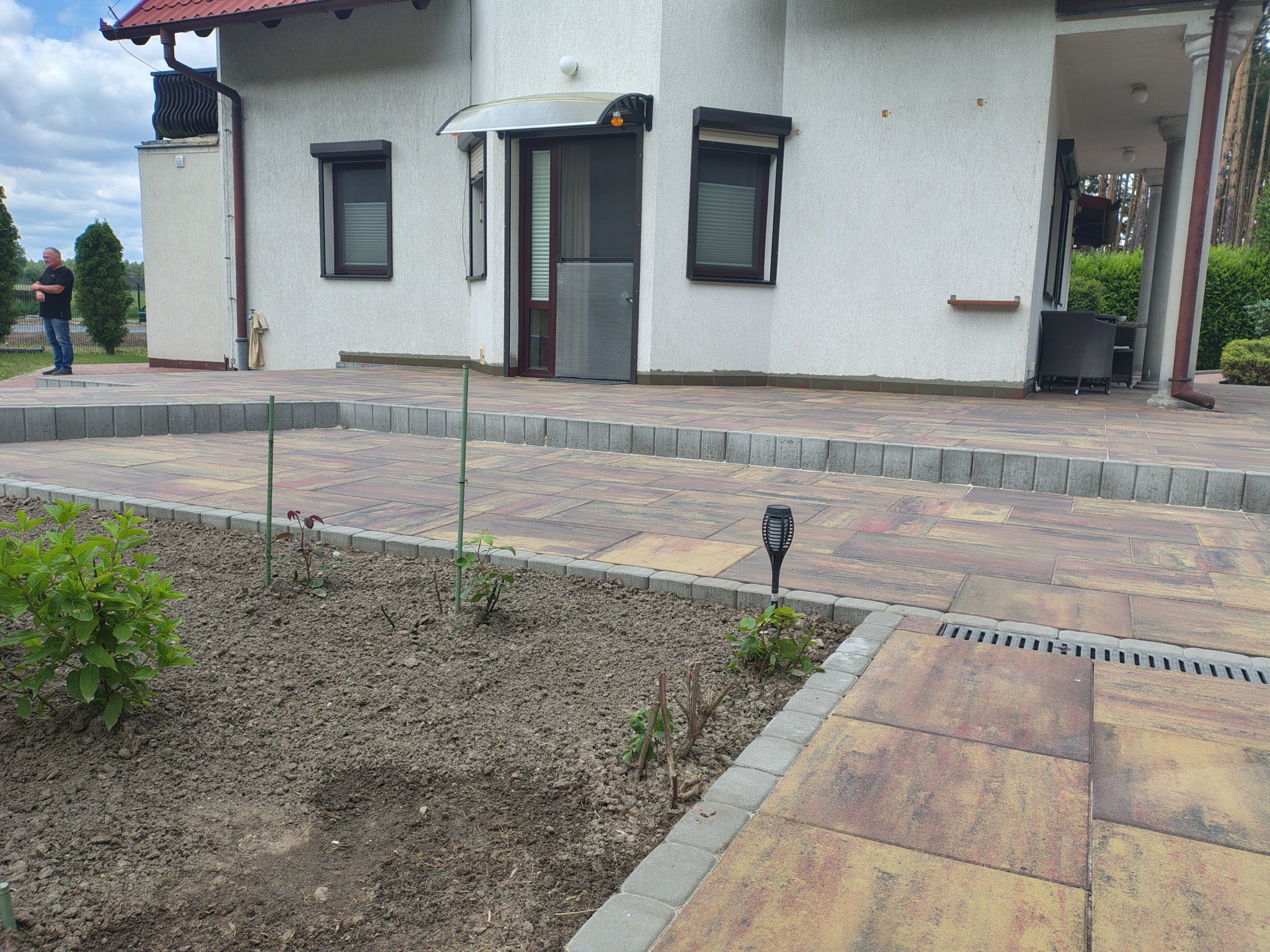 Ułożona kostka brukowa w odcieniach brązu i beżu przed domem z rabatami. Widoczny fragment elewacji z oknami i drzwi.
