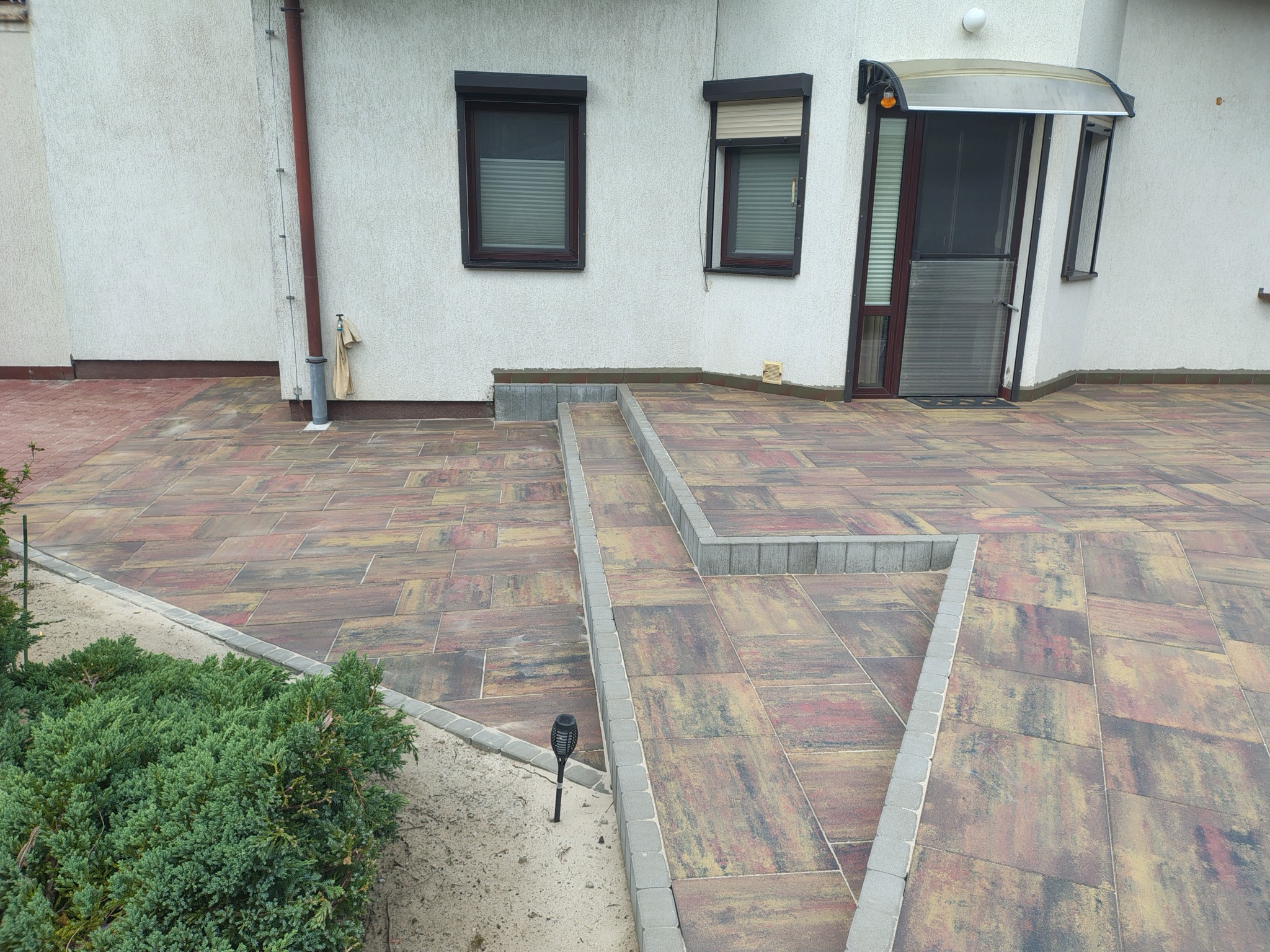 Kolorowa kostka brukowa tworzy ścieżkę z schodami, otoczoną szarą obwódką i zielonymi krzewami, prowadzącą do wejścia domu z białymi ścianami i brązowymi oknami.