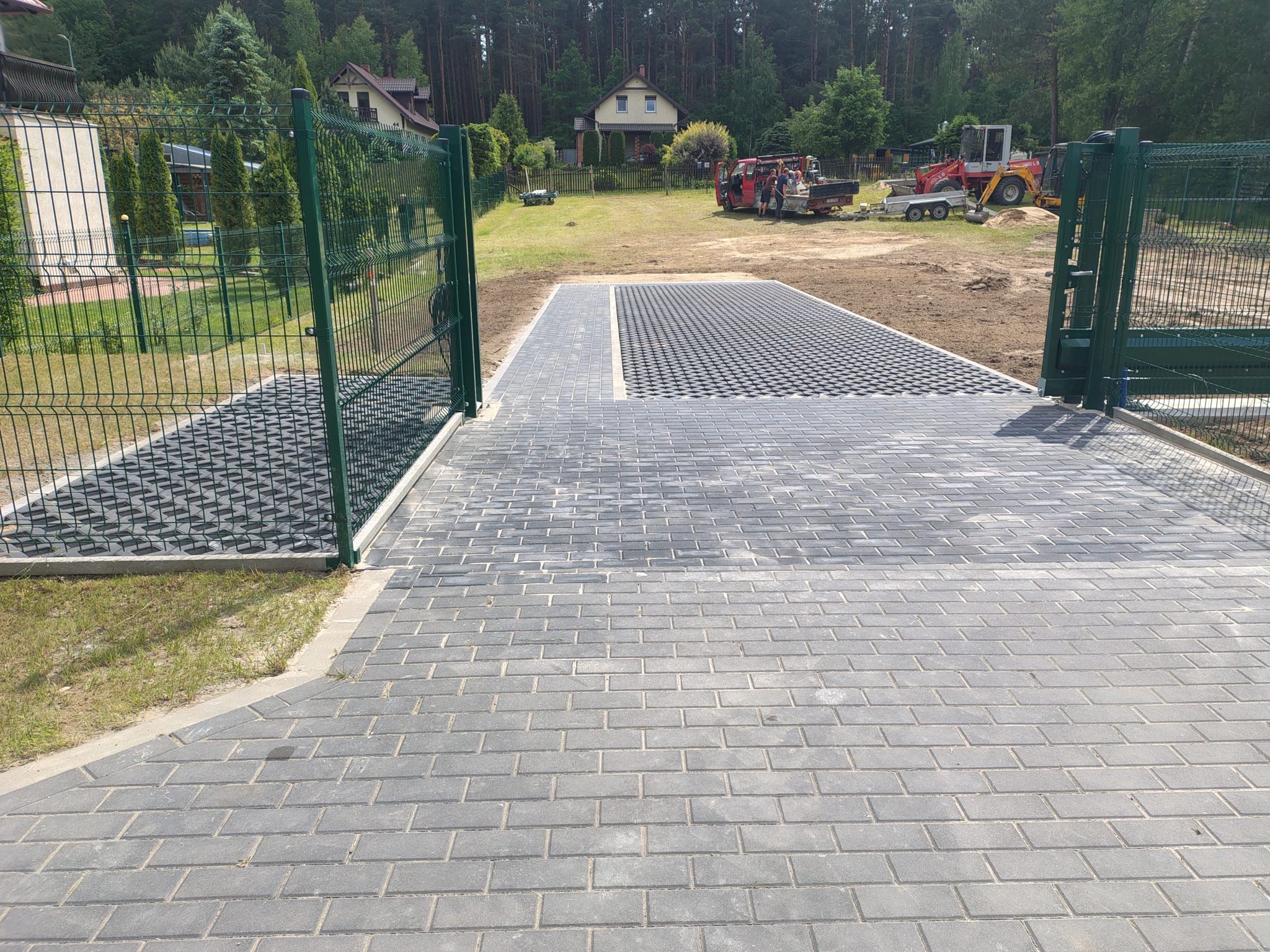Ułożona kostka brukowa i ażurowa przy wjeździe na posesję z zielonym ogrodzeniem panelowym. W tle dom i sprzęt budowlany.