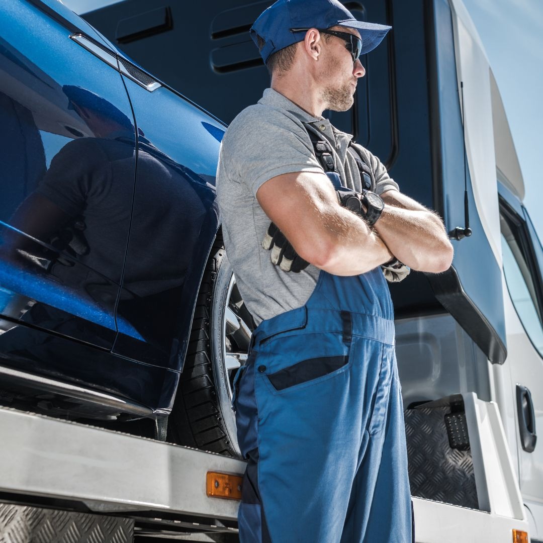 Mężczyzna w kombinezonie i czapce, z założonymi rękami, stoi obok niebieskiego samochodu na lawecie, w tle biała kabina lawety i błękitne niebo.