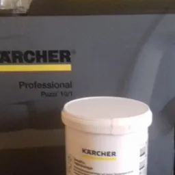 Profesjonalny odkurzacz piorący Kärcher Puzzi 10/1 z pojemnikiem na środek czyszczący Kärcher CarpetPro w jasnym otoczeniu.