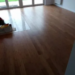 Ukończona podłoga z drewnianych paneli w jasnym pokoju z dużymi oknami balkonowymi i białym grzejnikiem na ścianie, widoczny fragment kartonu i płytek pod oknami.