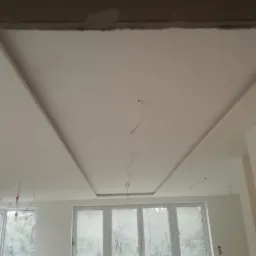 Biały sufit podwieszany z wnęką w kształcie trapezu, widoczne wystające przewody elektryczne, okno z widokiem na zewnątrz i fragment ściany z efektem marmuru.