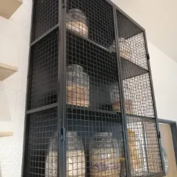 Metalowa szafka z siatki drucianej, zawieszona na ścianie, wypełniona słoikami z różnymi produktami spożywczymi, widoczna faktura ściany i fragment drzwi w tle.