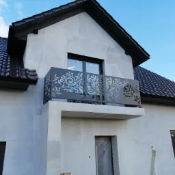 Nowoczesny balkon z ażurową, metalową balustradą o roślinnym wzorze, zamontowany w surowym stanie na budynku z szarą elewacją i ciemną dach&oacute;wką.