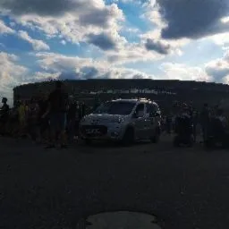 Szary minivan parkuje przed stadionem, otoczony przez grupę ludzi w słoneczny dzień.