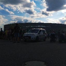 Szary minivan parkuje przed stadionem, otoczony przez grupę ludzi w słoneczny dzień.