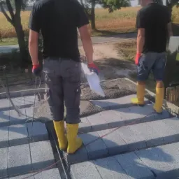 Dwóch pracowników w żółtych gumowcach stoi na stropie z szarych bloczków betonowych, zbrojonych prętami, widok z tyłu, w tle drzewa i pole.