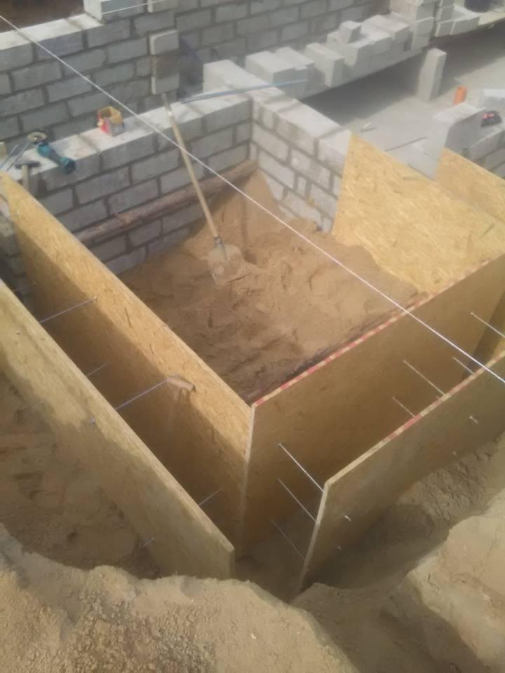 Wykop fundamentowy z drewnianym szalunkiem, wypełniony piaskiem i częściowo wymurowany z bloczków betonowych, widoczna łopata i narzędzia budowlane.