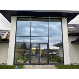Inwest-Profil Spółka z Ograniczoną Odpowiedzialnością Spółka Komandytowa - Duże, panoramiczne okno z przesuwnymi drzwiami, czarne ramy, widok na ogród, częściowo wykończony dom w tle, odbicie dwóch osób w szybie.