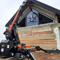 Inwest-Profil Spółka z Ograniczoną Odpowiedzialnością Spółka Komandytowa - Montaż okna o nietypowym trójkątnym kształcie przy użyciu specjalistycznego dźwigu z przyssawkami na placu budowy, widoczny pracownik w środku budynku.