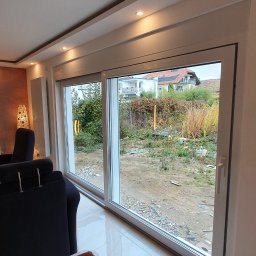 Inwest-Profil Spółka z Ograniczoną Odpowiedzialnością Spółka Komandytowa - Duże, przesuwne okno z białymi ramami, widok na zaniedbany ogród z chwastami i elementami wyposażenia, wnętrze z błyszczącą podłogą i fragmentem ciemnego fotela.