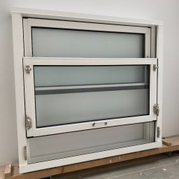 Inwest-Profil Spółka z Ograniczoną Odpowiedzialnością Spółka Komandytowa - Białe, przesuwne okno z dwoma skrzydłami, widoczne detale okuć i mechanizmów przesuwnych, umieszczone na drewnianych paletach, gotowe do montażu.