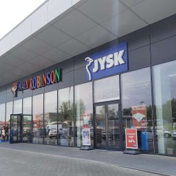 Inwest-Profil Spółka z Ograniczoną Odpowiedzialnością Spółka Komandytowa - Fasada nowoczesnego budynku handlowego z dużymi oknami wystawowymi, na których widoczne są plakaty reklamowe i informacje o wyprzedażach. Nad wejściami widoczne logotypy sklepów.