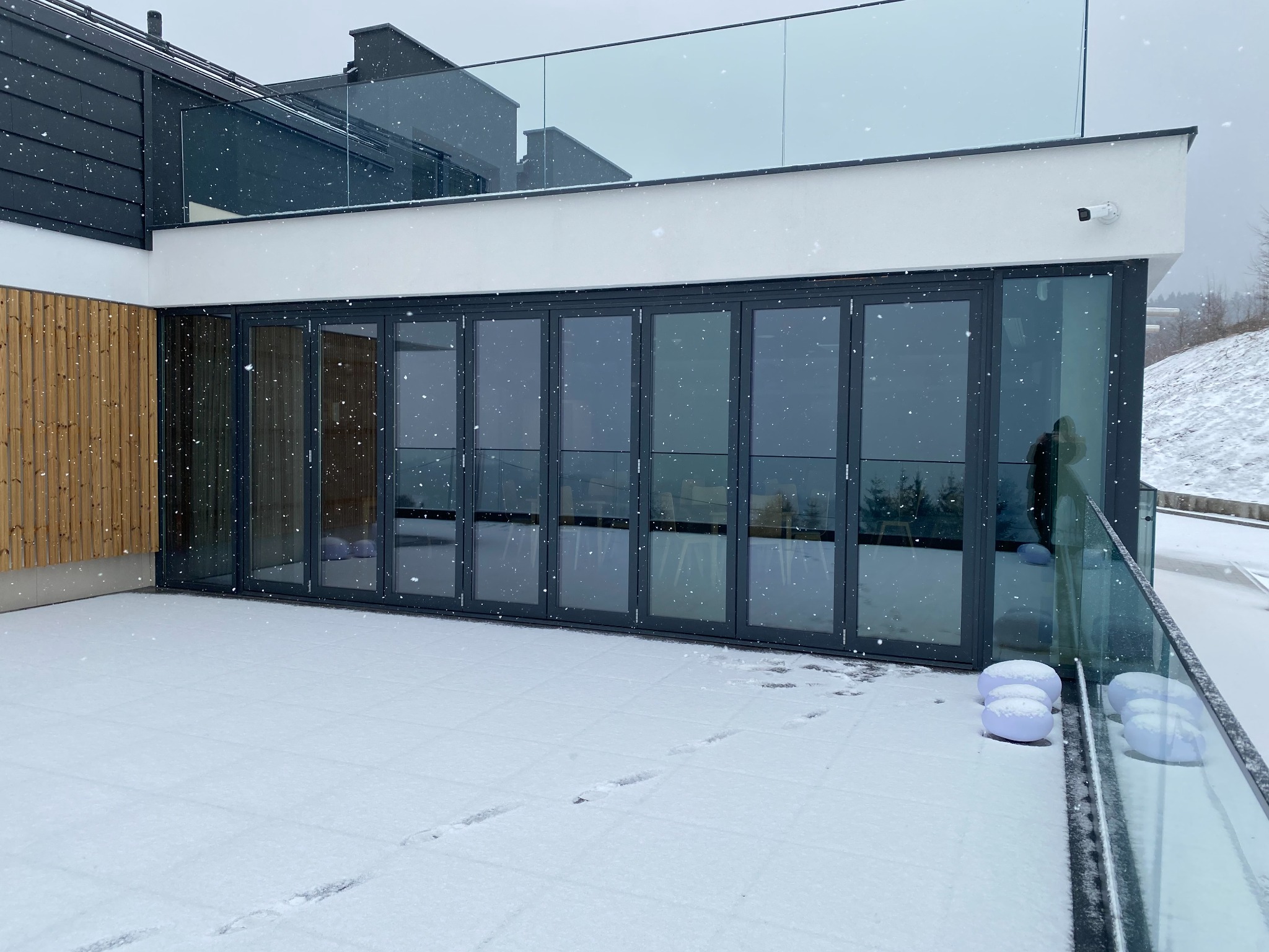 Zewnętrzne składane drzwi tarasowe w nowoczesnym domu, pokryte śniegiem, widoczne ślady stóp na tarasie, śnieg pada.