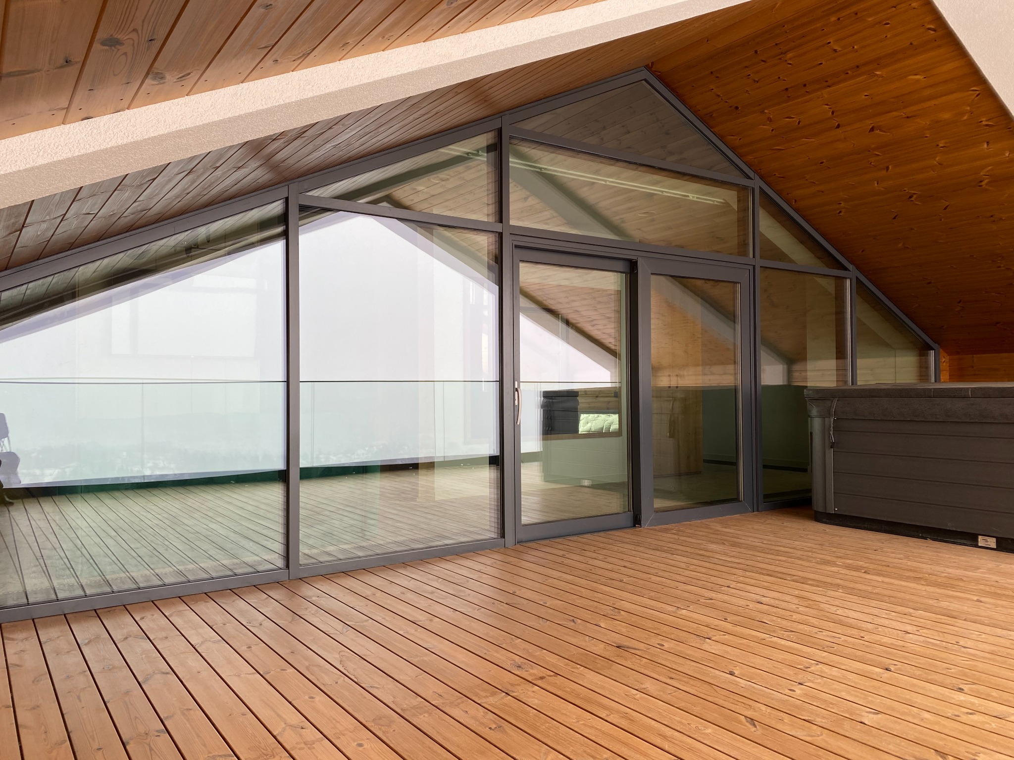 Zadaszony taras z drewnianą podłogą, przeszkloną ścianą z przesuwnymi drzwiami w grafitowych ramach i drewnianym sufitem pod skosem.