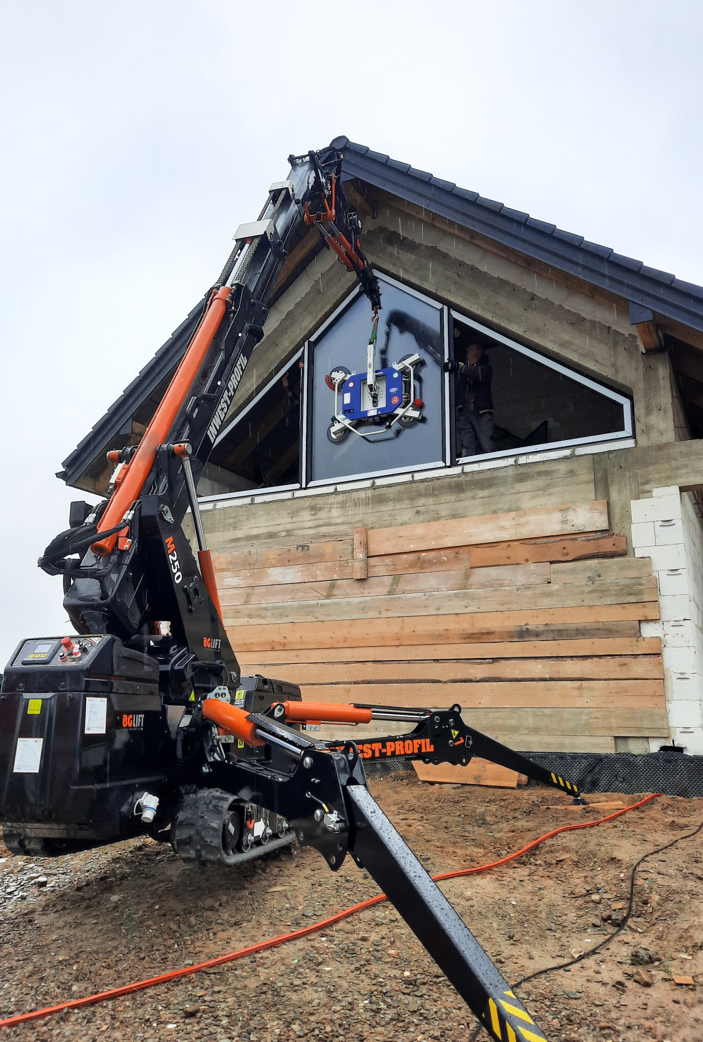 Montaż okna o nietypowym trójkątnym kształcie przy użyciu specjalistycznego dźwigu z przyssawkami na placu budowy, widoczny pracownik w środku budynku.