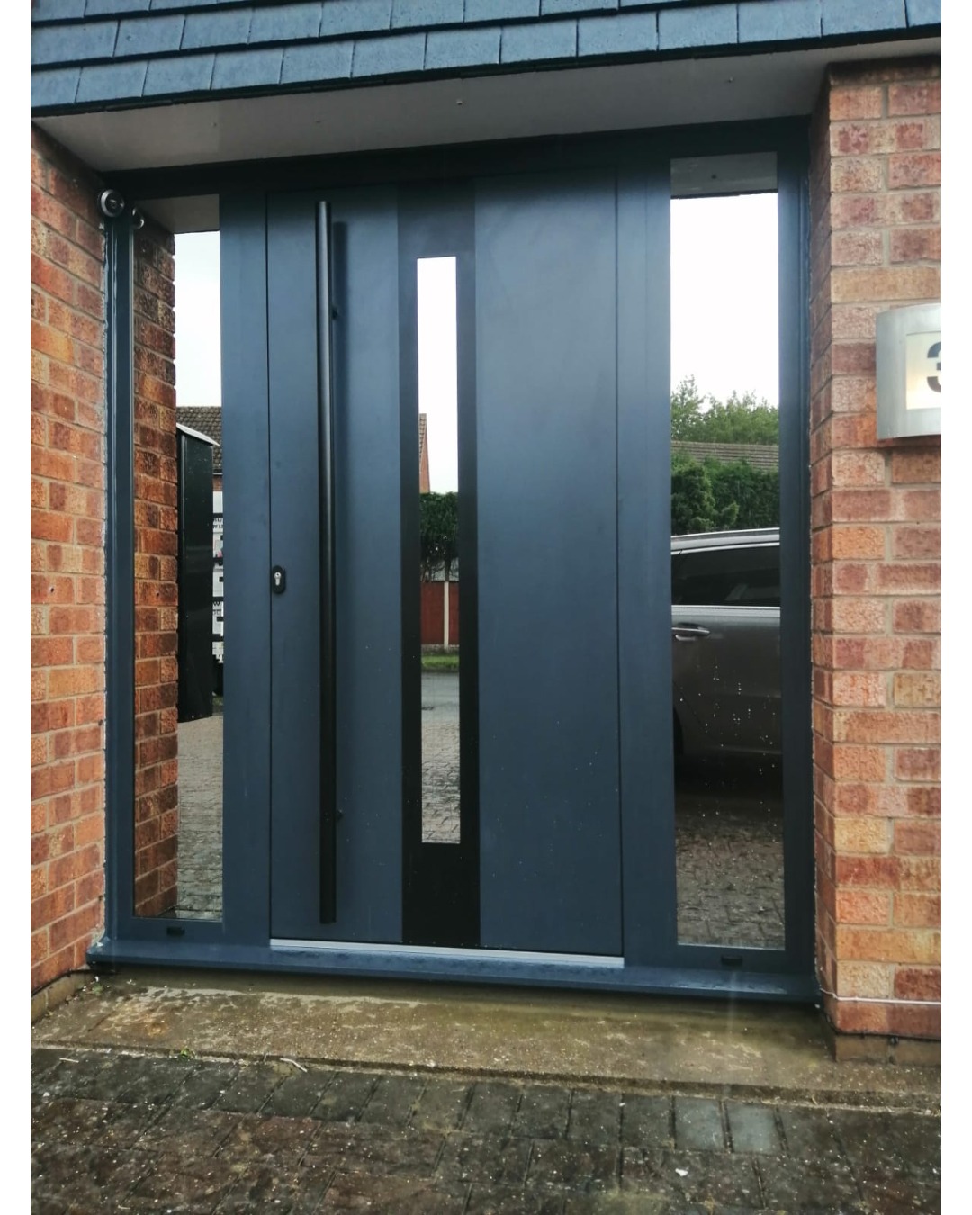 Nowoczesne, przesuwne drzwi wejściowe w kolorze grafitowym z długim, pionowym uchwytem i wąskim przeszkleniem, zamontowane w budynku z cegły.