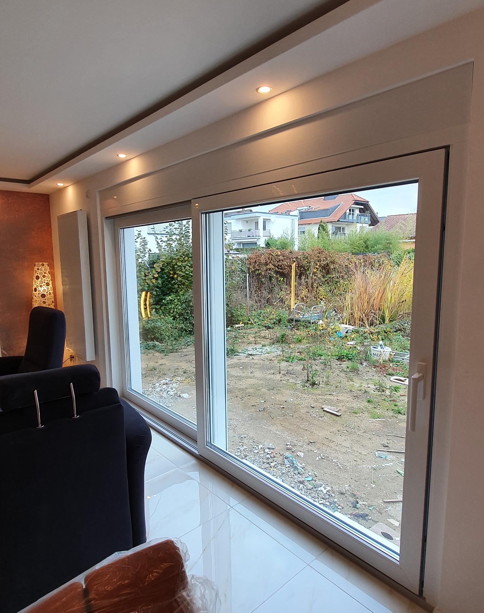 Duże, przesuwne okno z białymi ramami, widok na zaniedbany ogród z chwastami i elementami wyposażenia, wnętrze z błyszczącą podłogą i fragmentem ciemnego fotela.