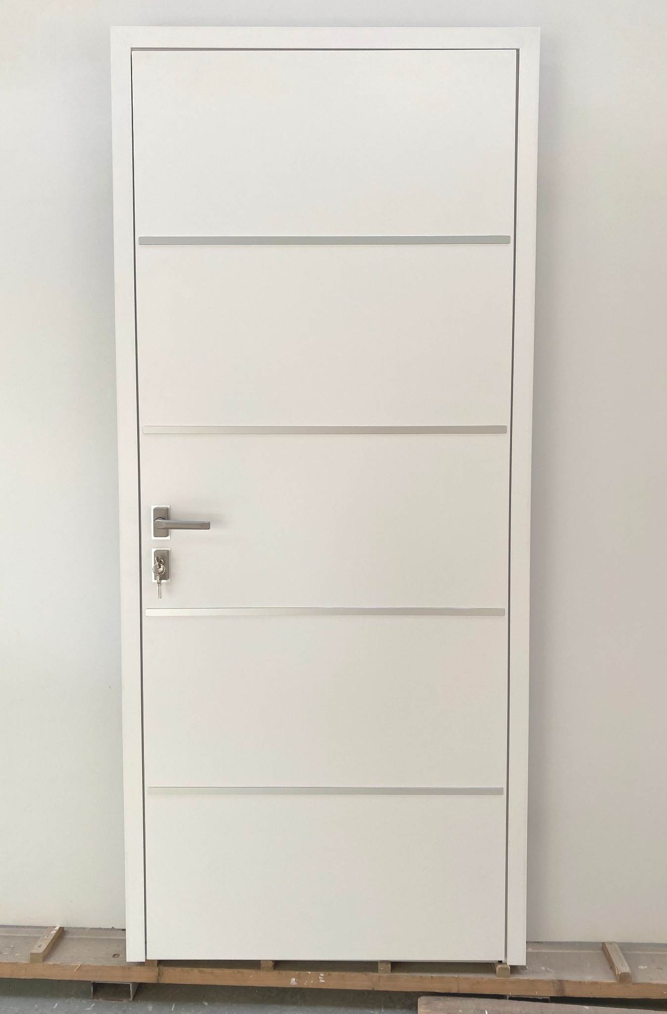 Białe drzwi wewnętrzne z poziomymi, srebrnymi wstawkami i klamką z kluczem, ustawione na drewnianej palecie, gotowe do montażu.