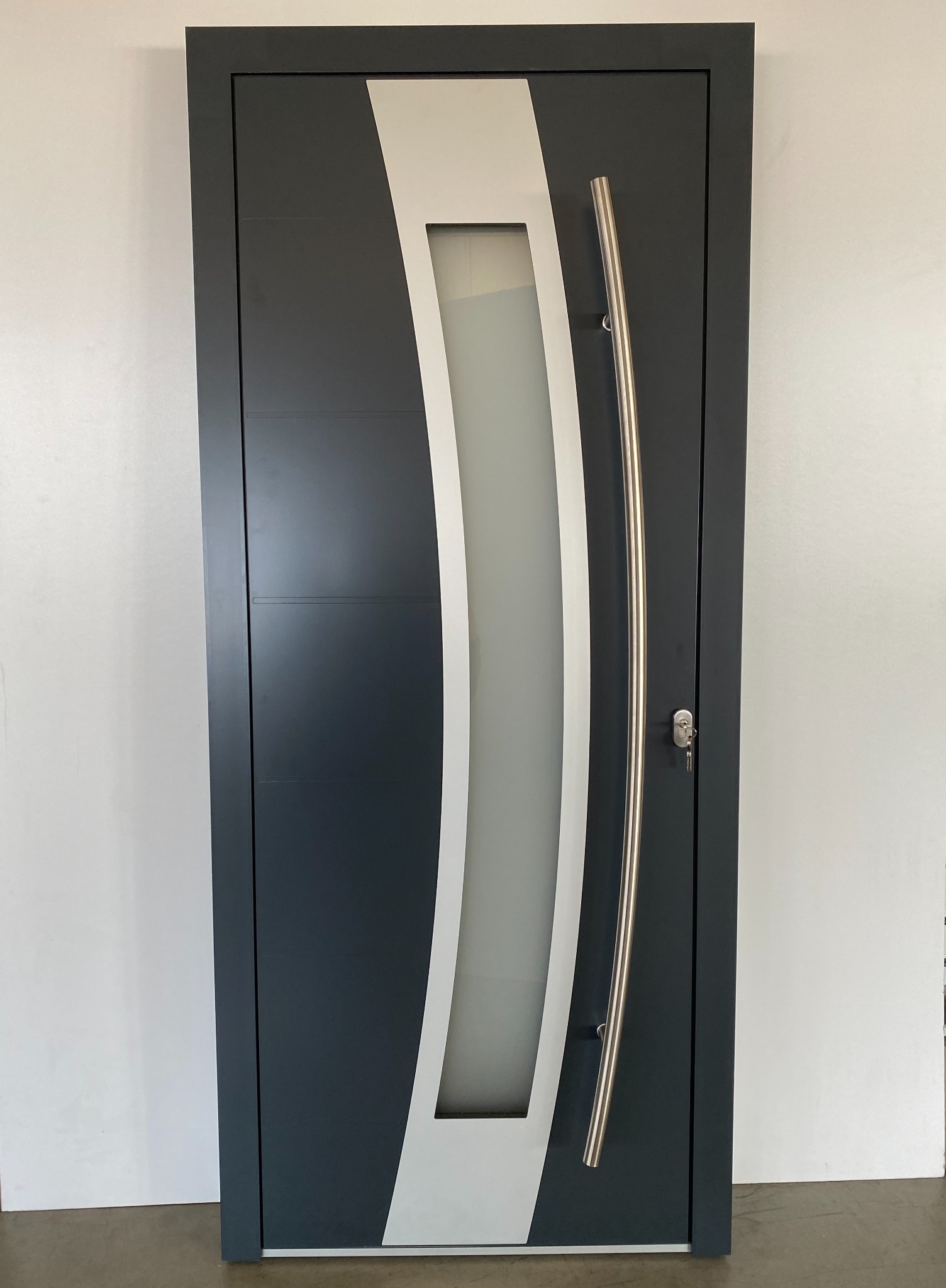 Nowoczesne drzwi wejściowe w kolorze grafitowym z długim, zakrzywionym uchwytem ze stali nierdzewnej i wstawką z mlecznego szkła, klucz w zamku.