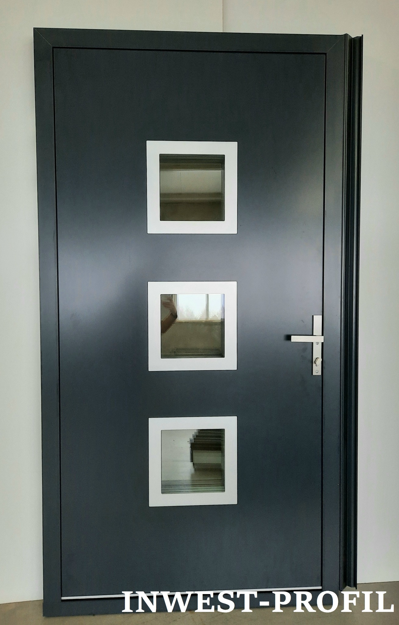 Nowoczesne, grafitowe drzwi z trzema kwadratowymi przeszkleniami w białych ramkach, widoczny srebrny uchwyt i logo firmy INWEST-PROFIL na dole.