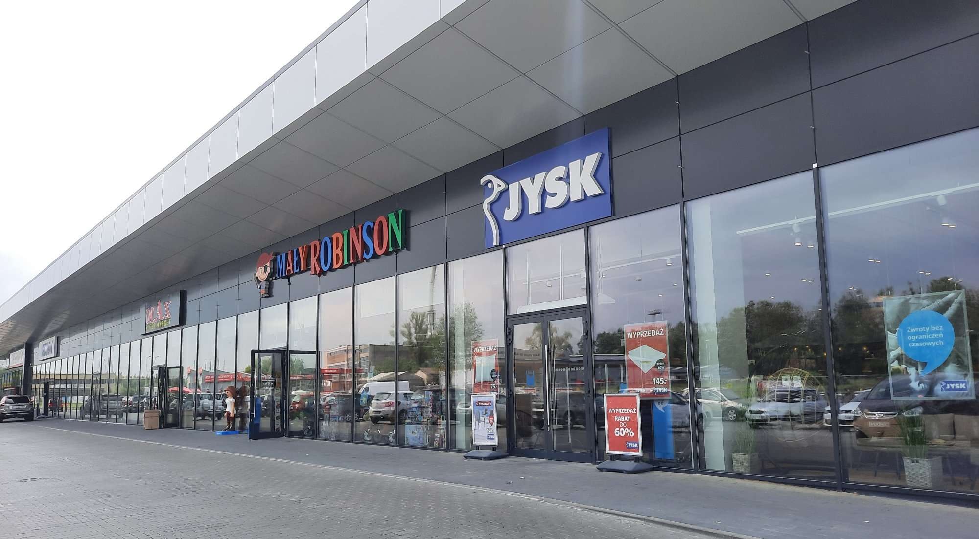 Fasada nowoczesnego budynku handlowego z dużymi oknami wystawowymi, na których widoczne są plakaty reklamowe i informacje o wyprzedażach. Nad wejściami widoczne logotypy sklepów.