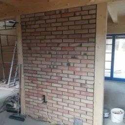 Ściana z cegły klinkierowej w trakcie montażu, widoczne przewody elektryczne wystające z otworu w ścianie, surowe drewniane elementy konstrukcyjne, drabina i narzędzia w tle.