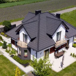 Nowoczesny dom jednorodzinny z ciemną dachówką, białymi ścianami i starannie zagospodarowanym ogrodem, widok z góry.