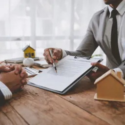 Mężczyzna w szarym garniturze wskazuje długopisem na dokument 'CONTRACT' na desce z klipsem, leżącej na drewnianym biurku z dwoma modelami domów i złożonymi rękami drugiej osoby w kadrze.