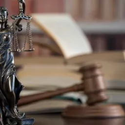 Statuetka Temidy z mieczem i wagą na tle sterty otwartych książek i drewnianego młotka sędziowskiego.