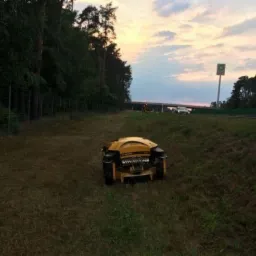 Żółty robot koszący trawnik na zboczu, w tle autostrada z wiaduktem i las.